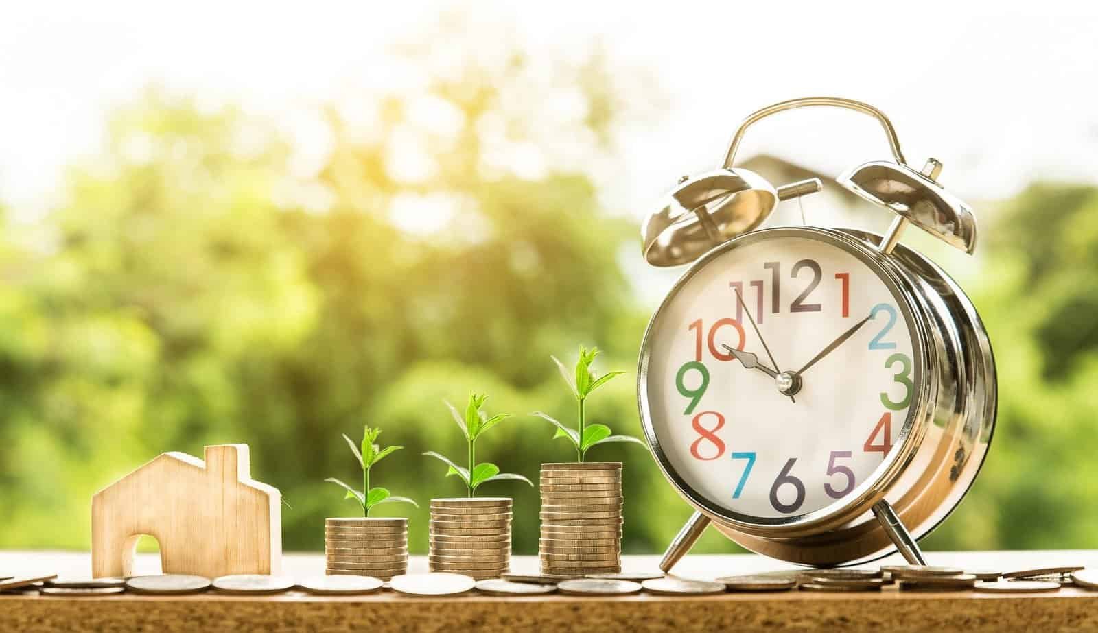 Real estate revenue growth concept with alarm clock, stacked coins with plants, and wooden house symbolizing time and investment returns